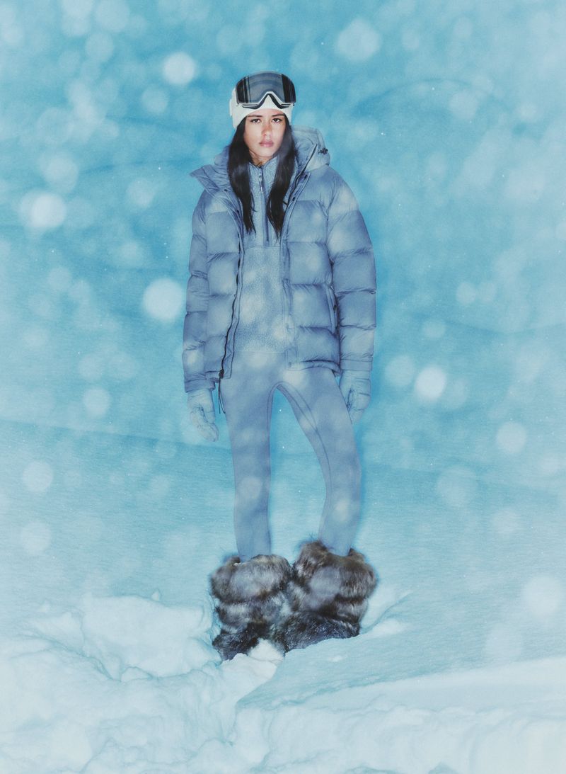 A woman stands in winter snow, wearing a ski outfit, puffer jacket, ski goggles, and fur boots.  Pale blue blurred background.