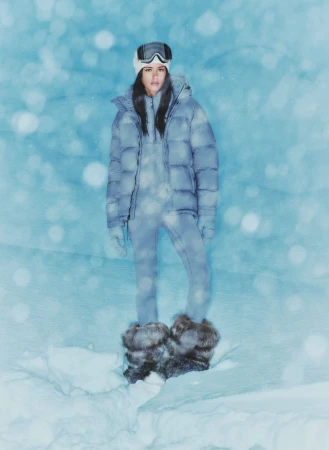 A woman stands in winter snow, wearing a ski outfit, puffer jacket, ski goggles, and fur boots.  Pale blue blurred background.