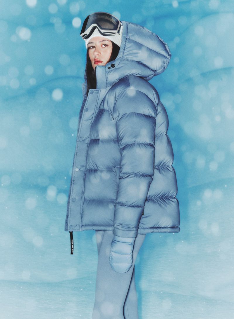 Woman in a powder blue puffer jacket with a hood and goggles poses in a snow-like environment with a light blue painted backdrop.