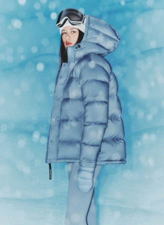 Woman in a powder blue puffer jacket with a hood and goggles poses in a snow-like environment with a light blue painted backdrop.