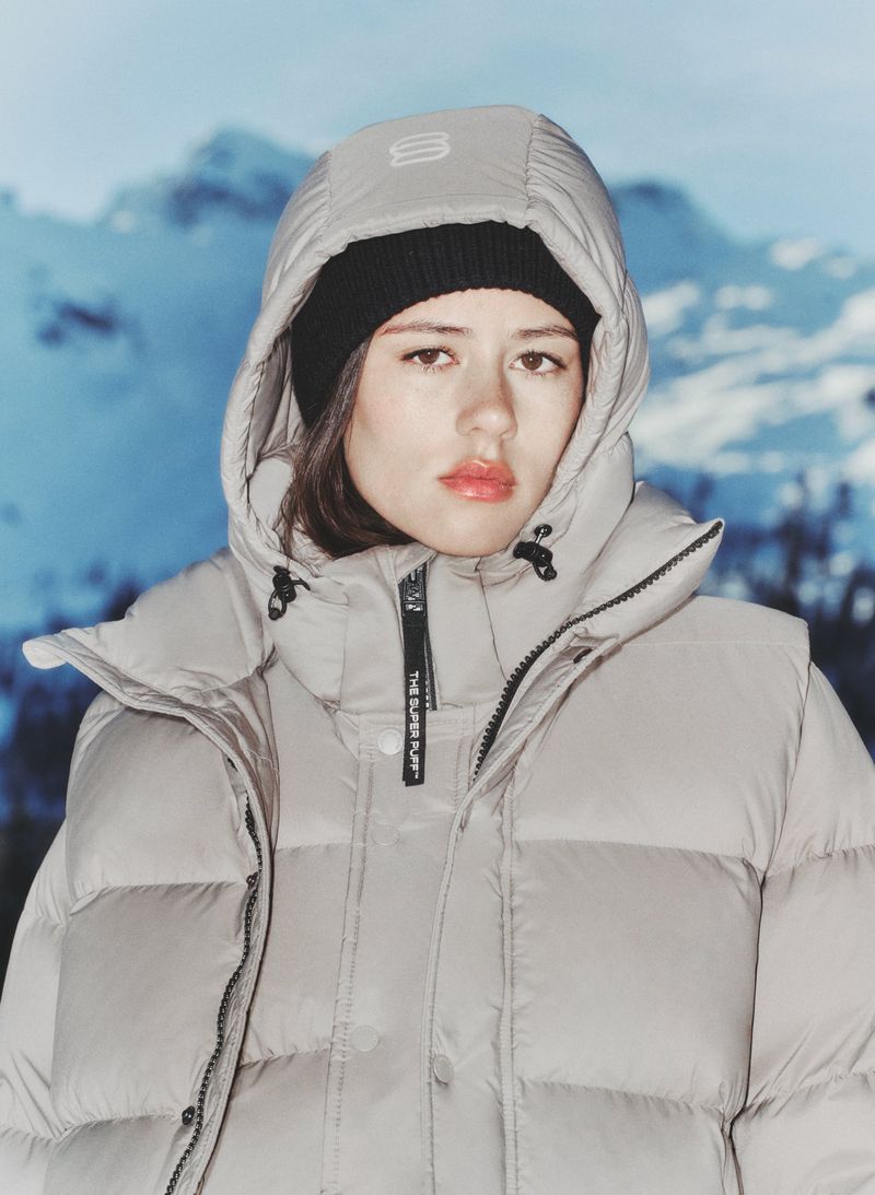 A young adult woman in a winter jacket and beanie poses in front of a blurred mountain background, highlighting winter fashion and outdoor apparel.