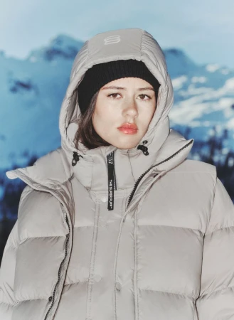 A young adult woman in a winter jacket and beanie poses in front of a blurred mountain background, highlighting winter fashion and outdoor apparel.