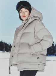 Woman wearing an oversized light grey puffer jacket and black ski goggles in a snowy outdoor landscape.