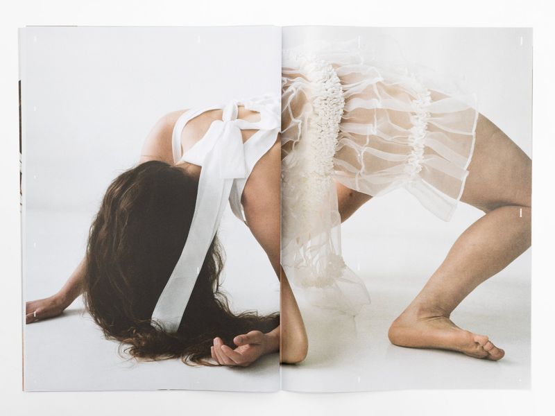 Artistic double-page magazine spread showcases a woman in flowing white fabric against a stark white background in studio lighting.