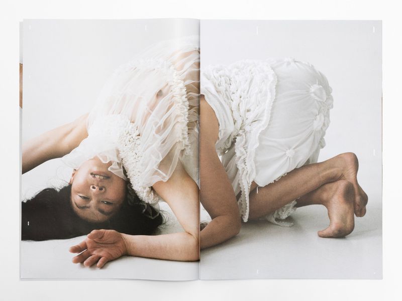 Fashion editorial shot of a woman in a sheer white dress kneeling on a white surface, captured with soft lighting and a dreamy aesthetic.