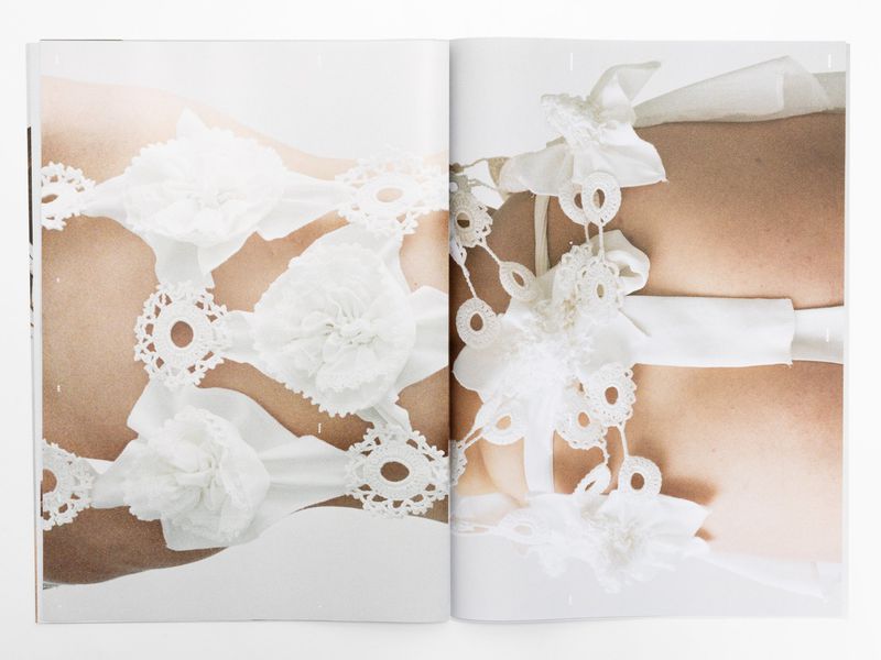 Detail of light-skinned torso adorned with delicate white lace, ribbons, and fabric flowers, set against a bright white background in a soft, diffused light.