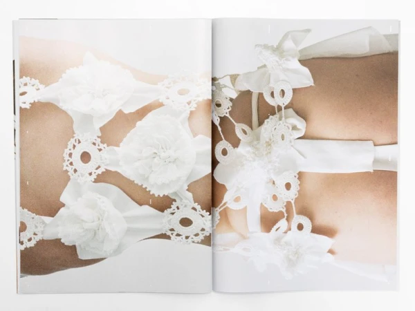 Detail of light-skinned torso adorned with delicate white lace, ribbons, and fabric flowers, set against a bright white background in a soft, diffused light.
