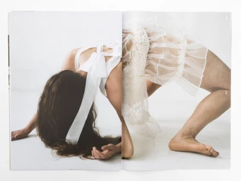 Model in an arched backbend pose wearing a white ruffled sheer garment with a back bow against a white backdrop.