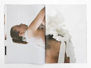 Open editorial publication spread featuring fashion photography of a model wearing delicate white fabric and a ruffled headpiece.