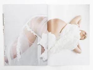 Model lying down wearing textured sheer white sculptural garments against a bright white background in an open editorial spread.