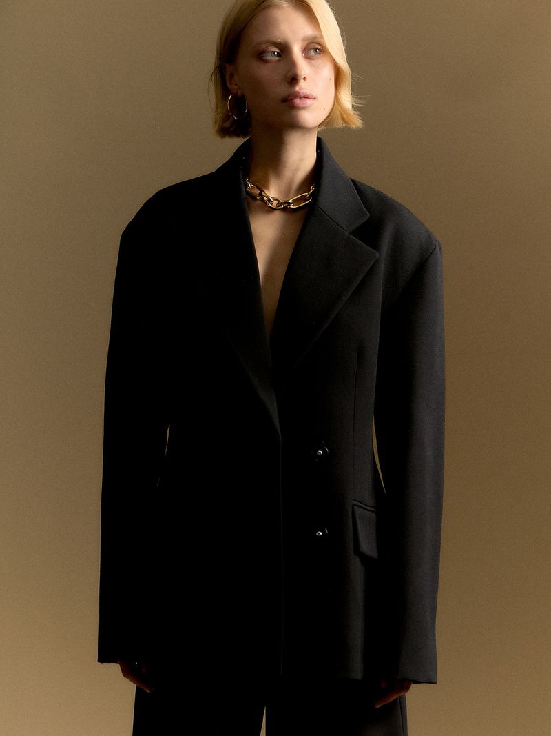 A blonde woman in a black blazer stands in front of a beige background, accessorized with gold hoop earrings and a gold necklace.