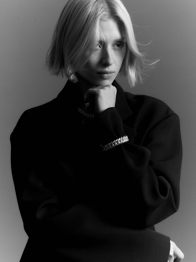 Monochrome portrait of a young woman with blonde hair in a black blazer, resting her chin on her hand, accessorized with jewelry.