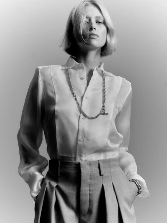 Black and white portrait of a fair-skinned model wearing a white collared shirt, grey pants, and a silver chain necklace in a studio.