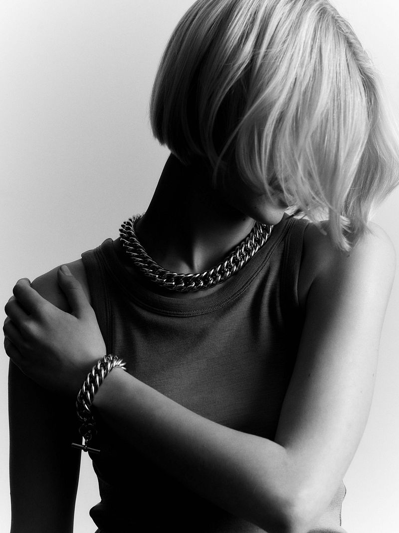 Monochrome portrait of a blonde woman wearing a chain necklace and bracelet, set against a bright white background in a photo studio.