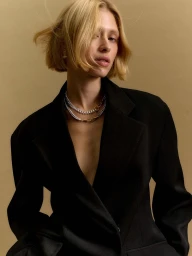 Model with blonde bob wearing a black blazer and layered chain necklaces against a neutral tan background.