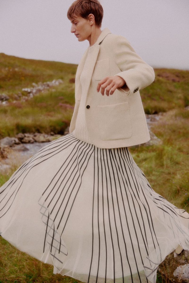 Adult woman walks in a field wearing a beige jacket, cream sweater, and long flowing striped skirt in an outdoor landscape setting.