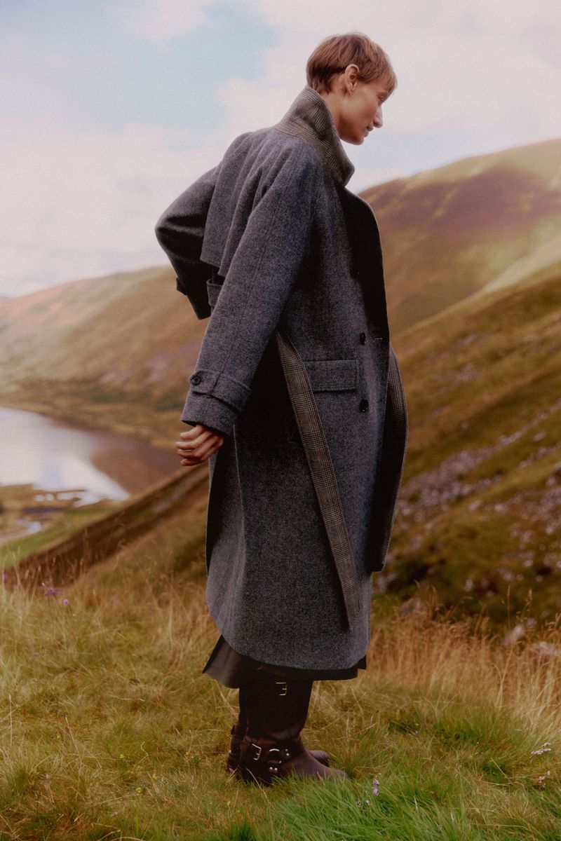 Model wearing a long grey coat standing in a grassy mountain landscape overlooking water for ME+EM