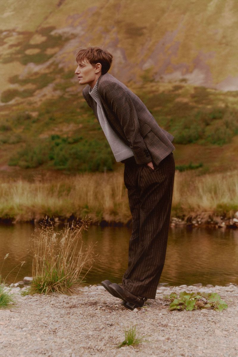 Adult in a tweed suit bending by a river surrounded by mountains and greenery. Natural light and earth tones lend to the image's mood.