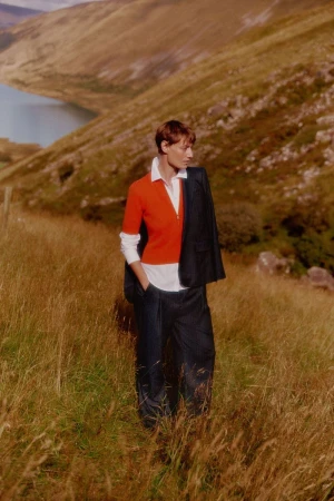 Fashionable adult poses in a grassy landscape with a lake and mountains. They wear a red top, blazer, and pants for a stylish shot.