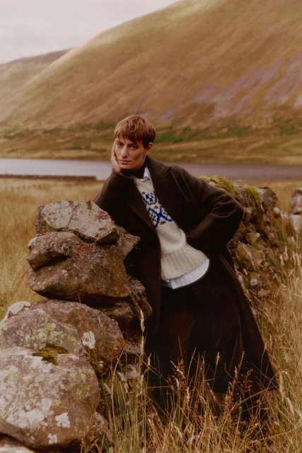 Adult woman in a coat and sweater leans against a stone wall in a grassy rural landscape with a mountain range backdrop.