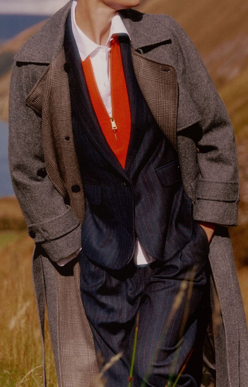 Model in tweed coat, navy suit, and orange top in a grassy field with distant lake and hills, fashion editorial outdoor shot.