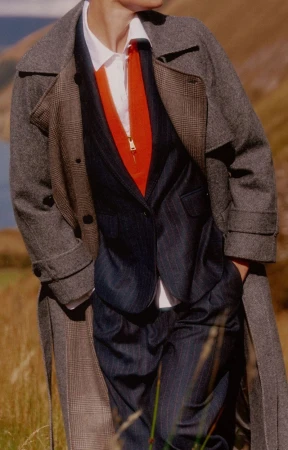 Model in tweed coat, navy suit, and orange top in a grassy field with distant lake and hills, fashion editorial outdoor shot.
