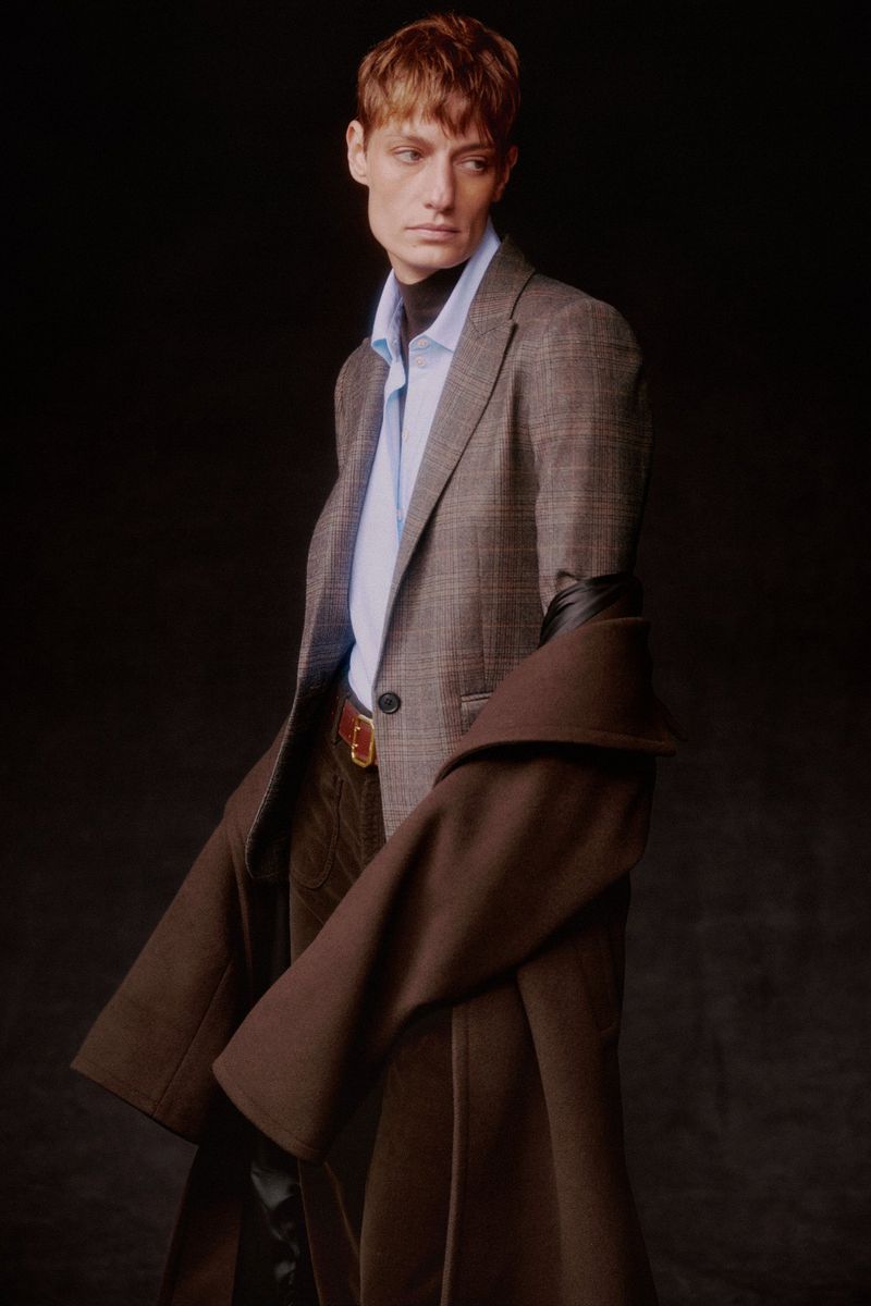 Male model poses in a brown suit and trench coat against a dark background, wearing a light blue shirt and brown belt.