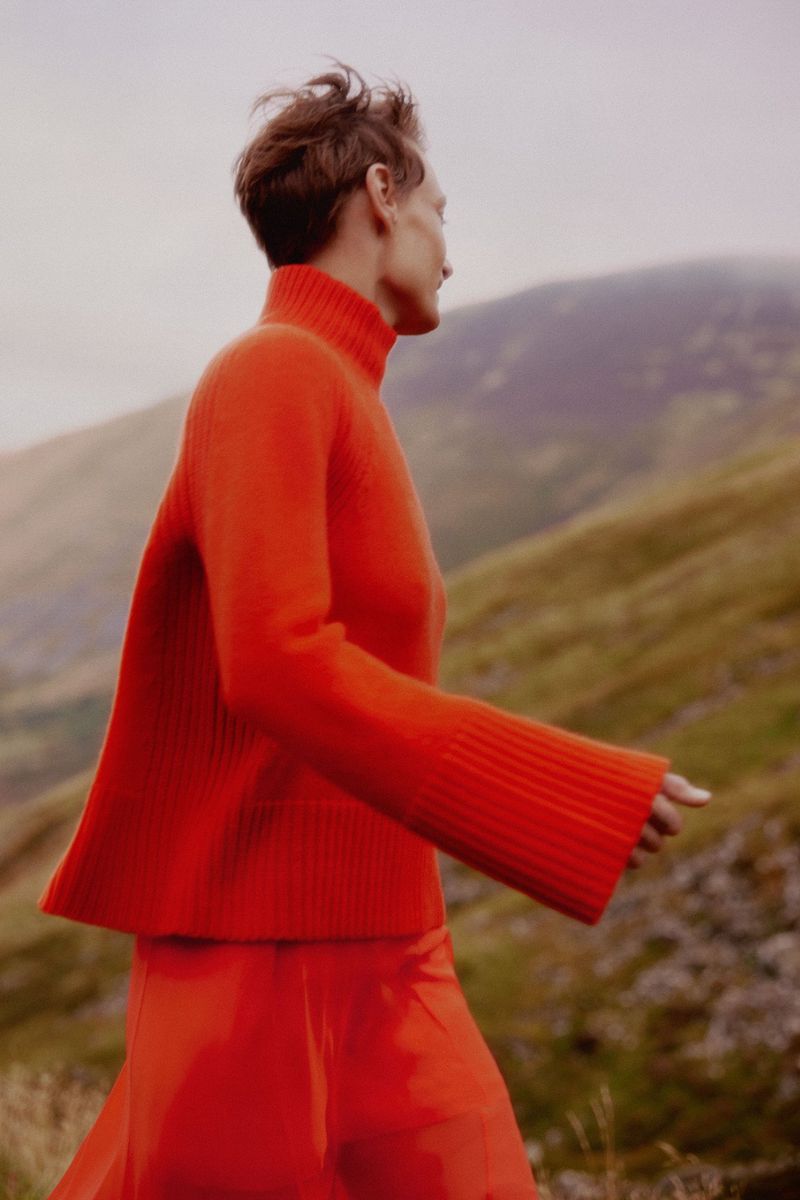 Woman in bright orange turtleneck sweater and skirt walks across a grassy hillside with mountains in the distance on an overcast day.