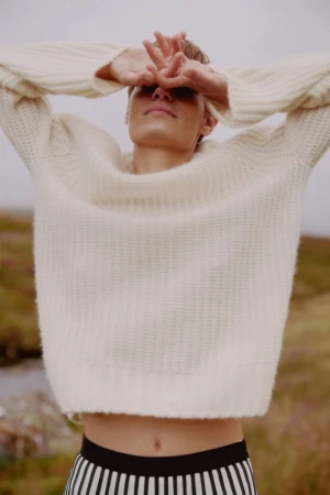 Model wearing a textured cream sweater with arms raised, covering eyes against a muted landscape background