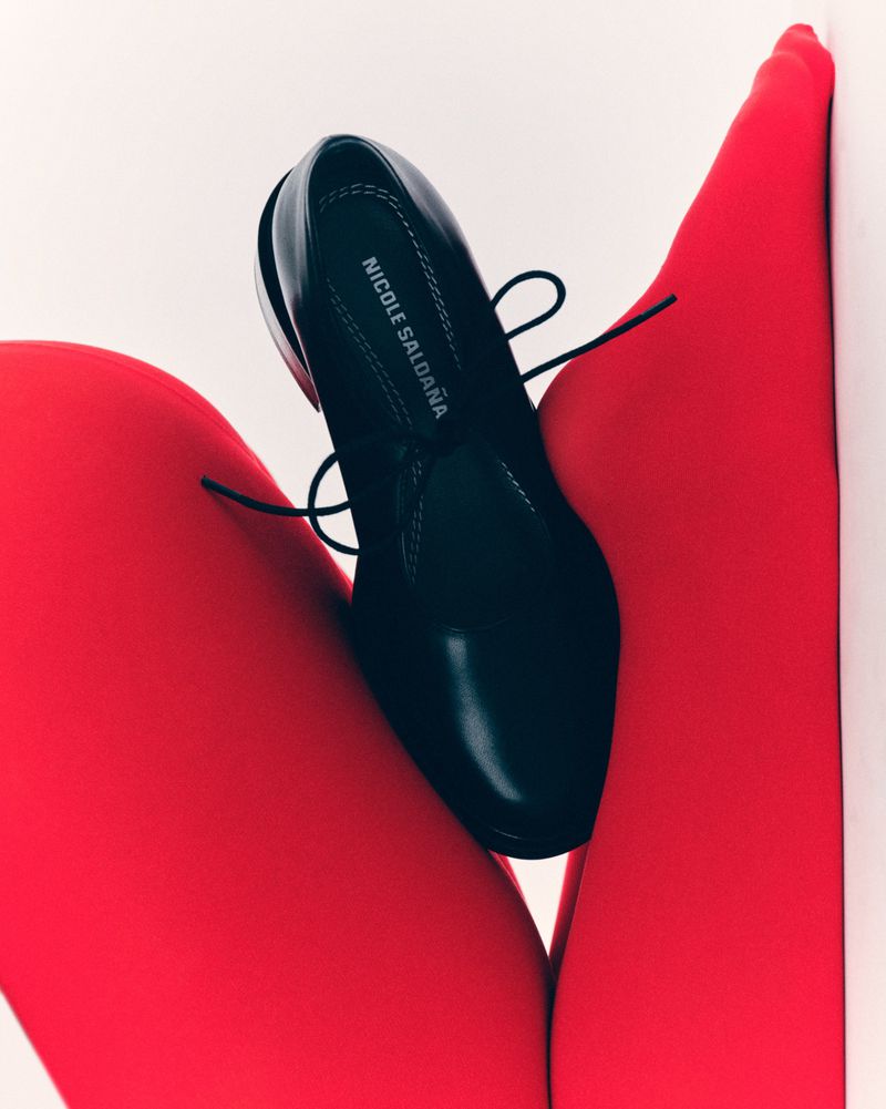 Studio shot of a black leather dress shoe by Nicole Saldana on a pair of red tights against a white background.