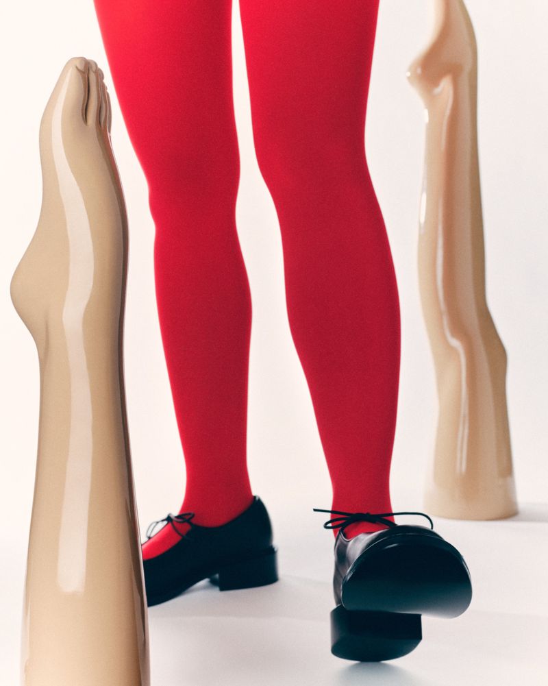 Close-up fashion shot of person wearing red tights and black shoes standing between two mannequin legs against a white background.