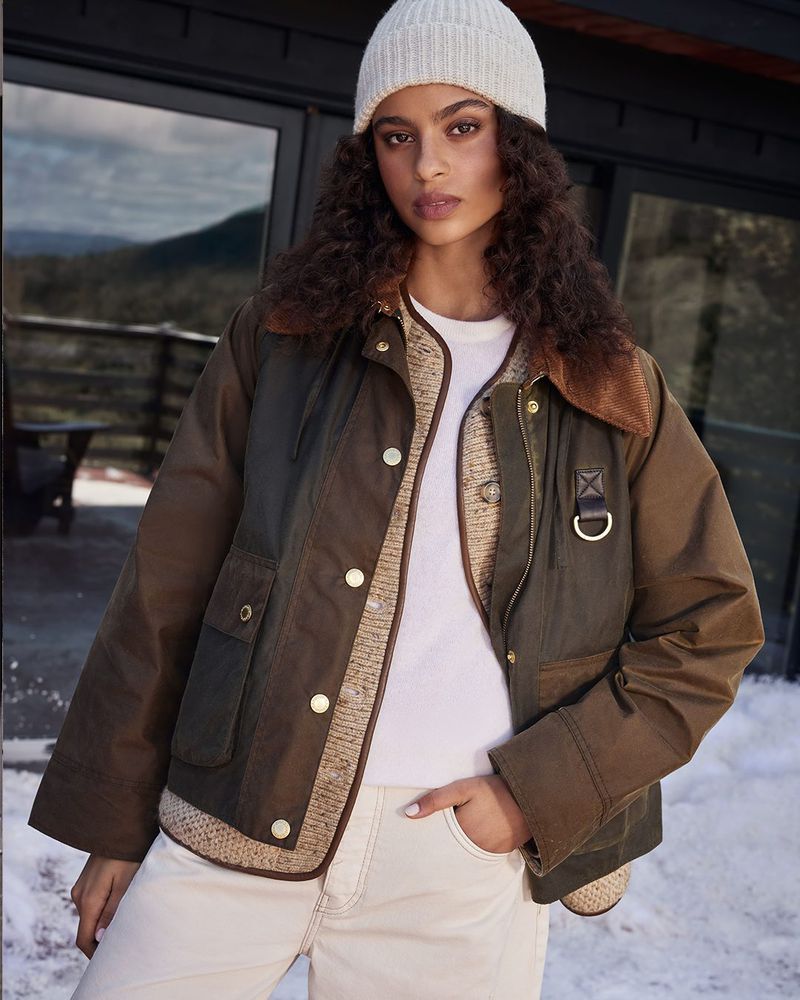 A woman with curly hair wearing a knit hat and a brown jacket poses outdoors against a backdrop of snowy hills and a cabin.
