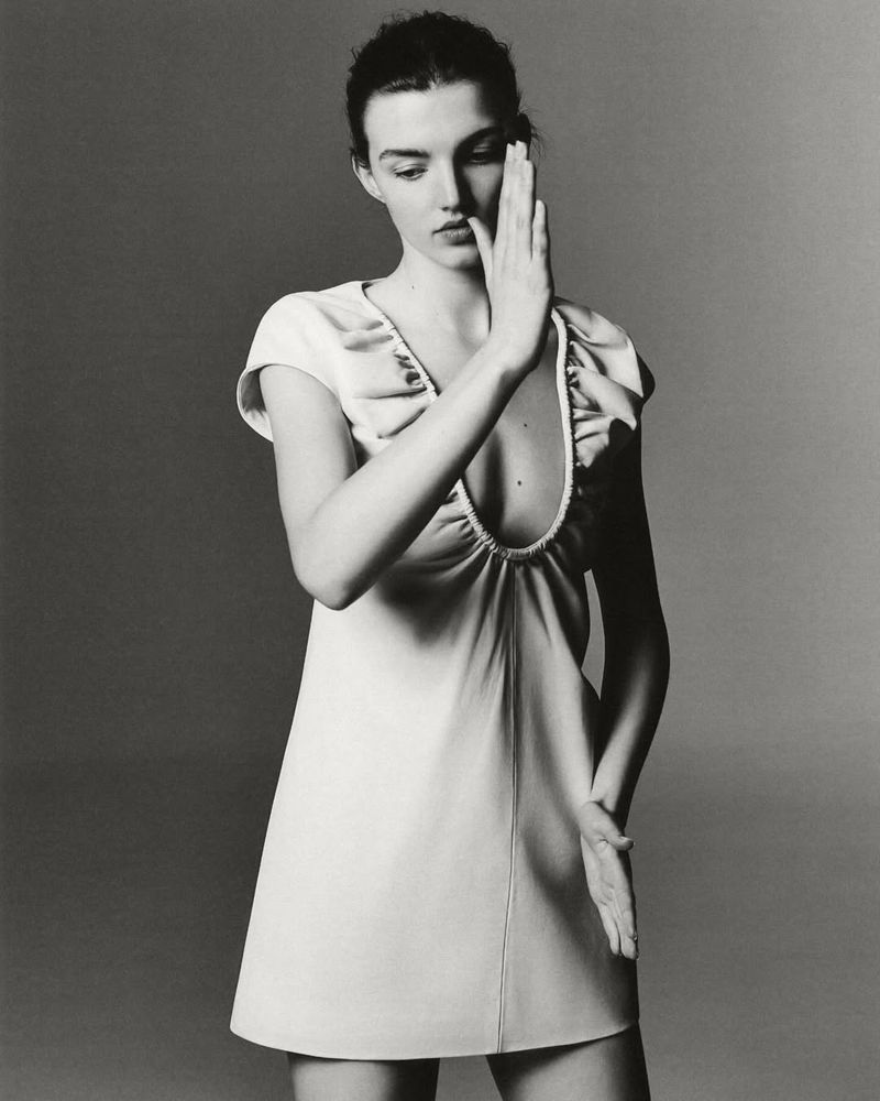 Monochrome studio portrait of an adult woman with her hands up, wearing a cream-colored A-line dress with a low neckline and short sleeves.