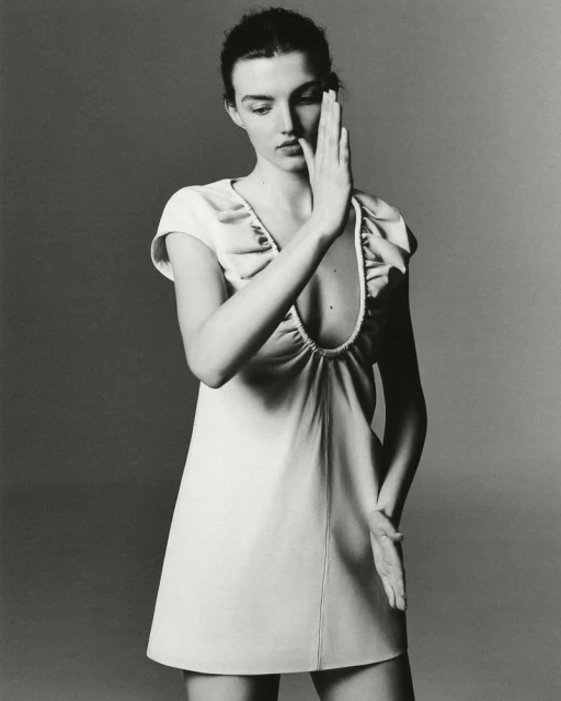 Monochrome studio portrait of an adult woman with her hands up, wearing a cream-colored A-line dress with a low neckline and short sleeves.