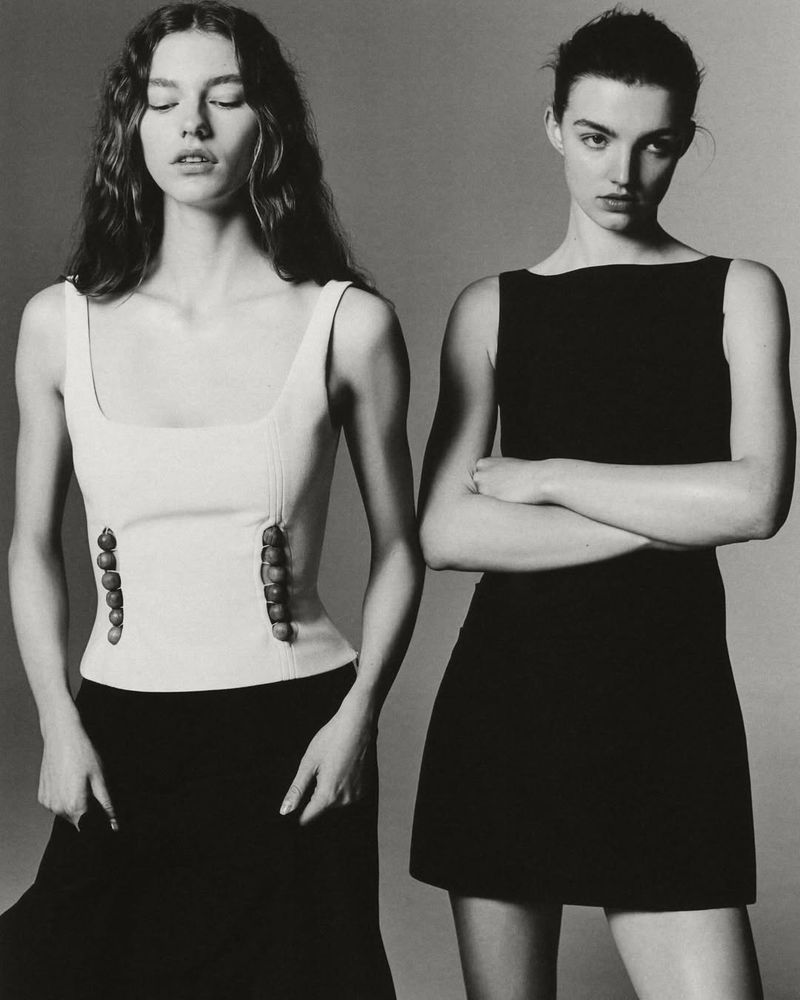 Two women in black and white photograph posing in dresses. One woman has her arms crossed and the other has her arms down.
