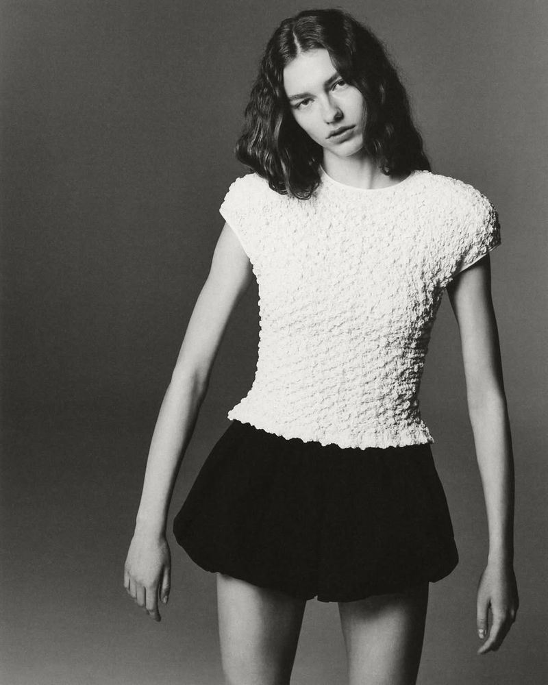Monochromatic portrait of a young adult female fashion model posing indoors, wearing a white textured top and black shorts.