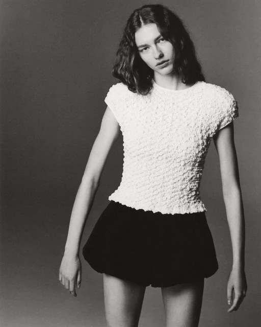 Monochromatic portrait of a young adult female fashion model posing indoors, wearing a white textured top and black shorts.