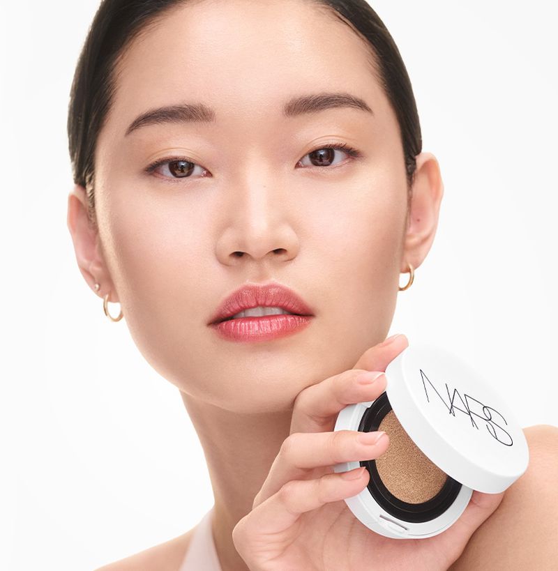 Close-up of an Asian woman holding a NARS foundation compact. The model is fair-skinned with her hair pulled back, simple gold hoop earrings.