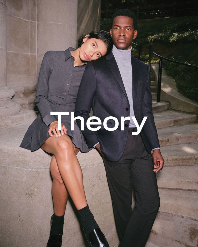 A diverse couple posing outdoors; the woman in a dress and loafers leans on the shoulder of the man in a blazer and trousers.
