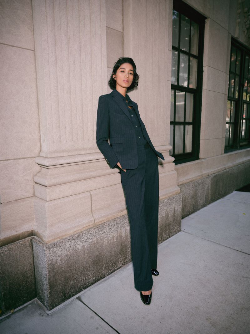 Woman in a dark pinstripe suit standing next to stone columns. Dark blazer and wide-legged trousers, dark hair, black shoes.