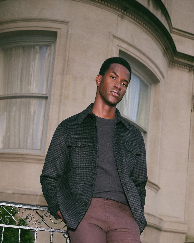 An adult man leans against a wrought iron railing outdoors, posing in a charcoal shirt jacket over a t-shirt and brown trousers.