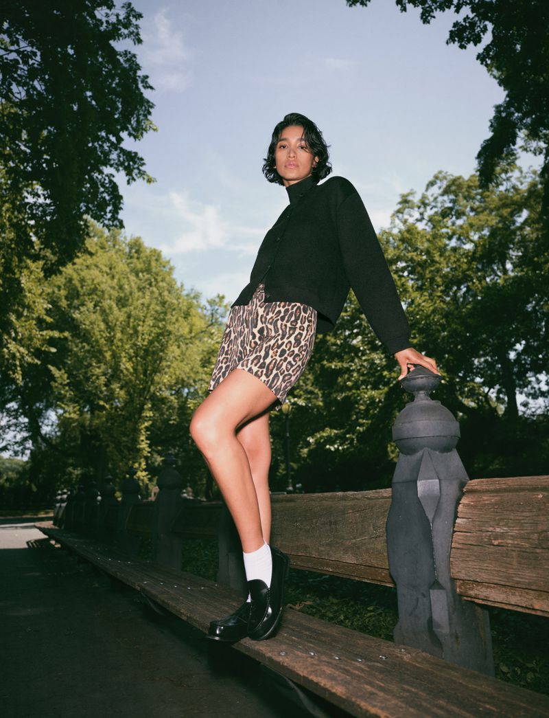 Woman in black jacket and animal print shorts standing on park bench in dappled light. Trees and blue sky visible in background.