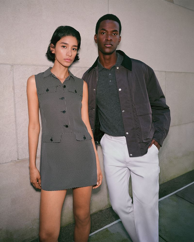 Two fashion models, an adult woman in a patterned minidress and an adult man in a jacket and pants, pose against a wall in a studio.