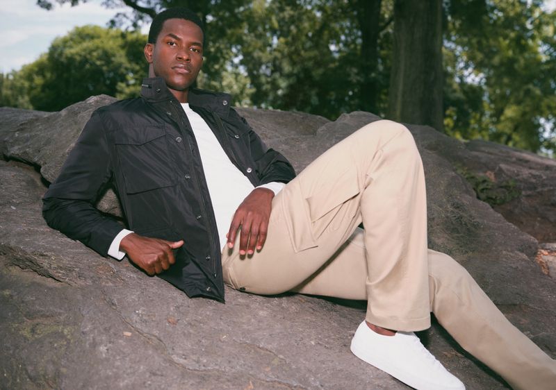 Man in black jacket poses on a rock outdoors in a natural setting wearing tan pants and white sneakers with green trees in background.