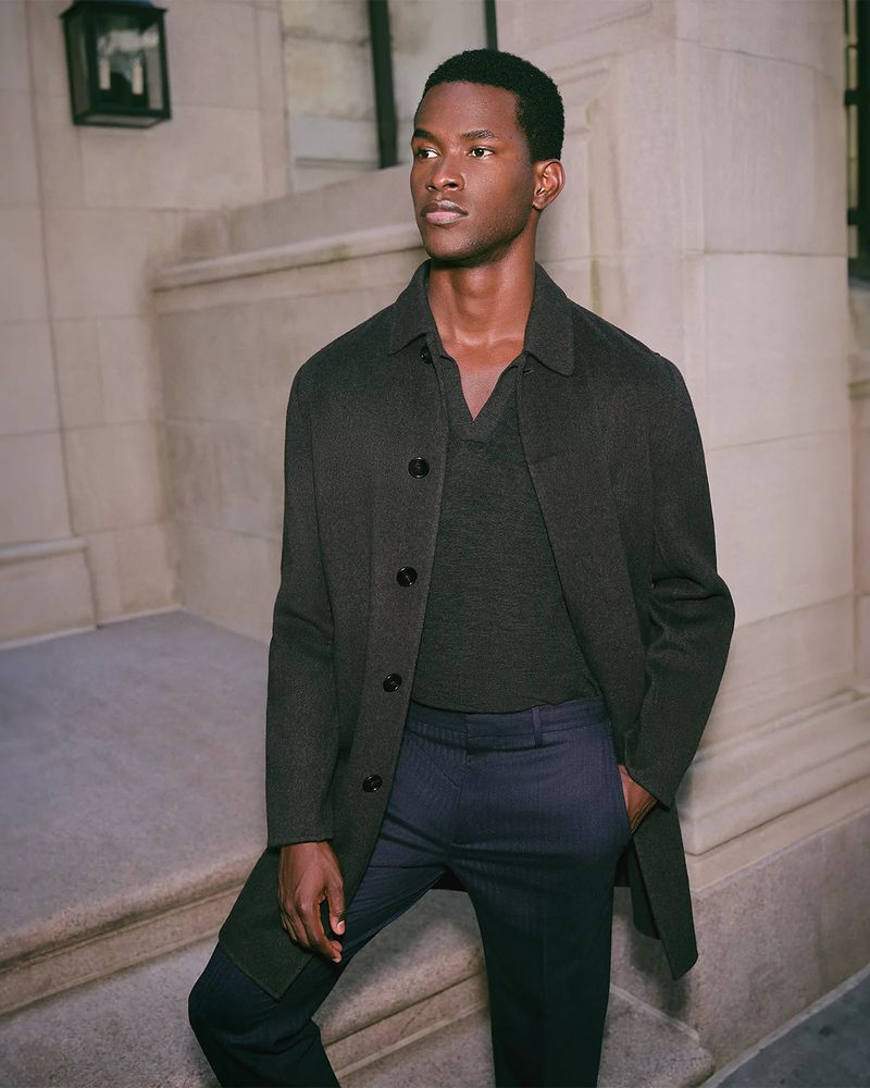 Man in dark overcoat and v-neck sweater stands on stairs, styled in dark blue dress pants against a textured, neutral background.