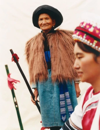 Elderly Asian woman in traditional dress with a fur shawl and black hat, holding a wooden staff, outdoor against a light background.