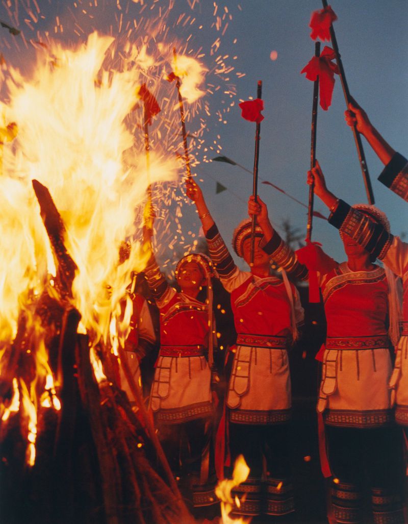 Group of people in traditional dress holding ceremonial staffs with red cloth, performing ritual near large bonfire against dusk sky.