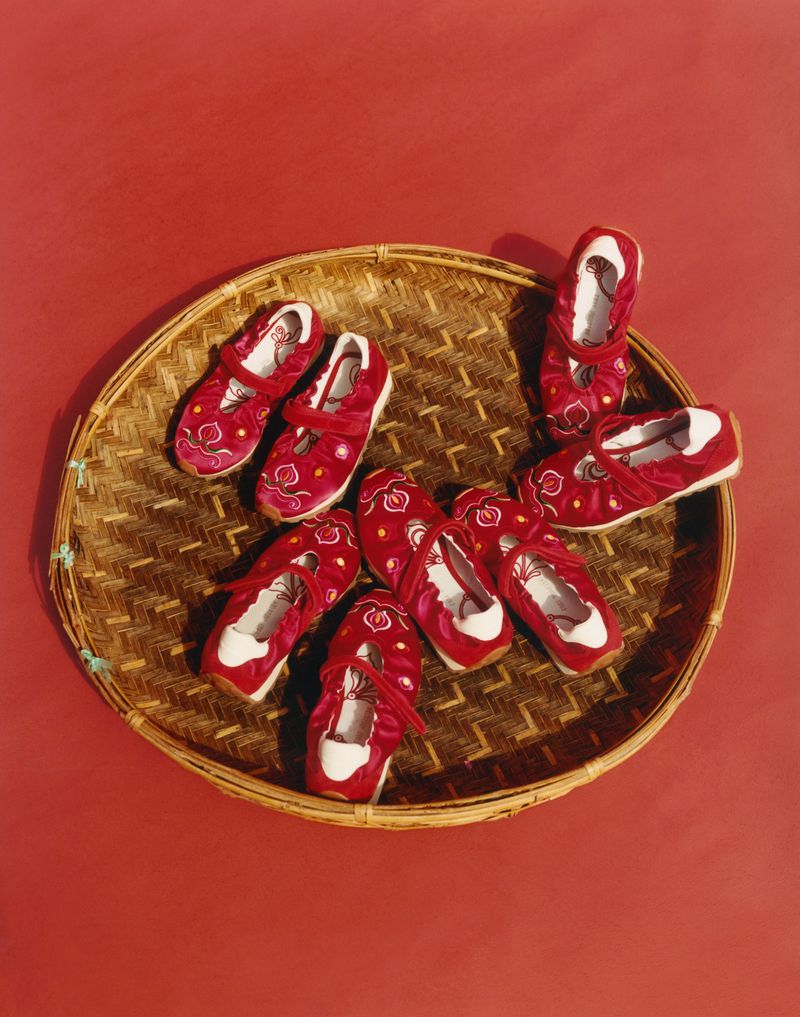 Overhead shot of traditional Chinese children's shoes with red velvet, white trim, and embroidery, arranged on a woven basket against a red backdrop.