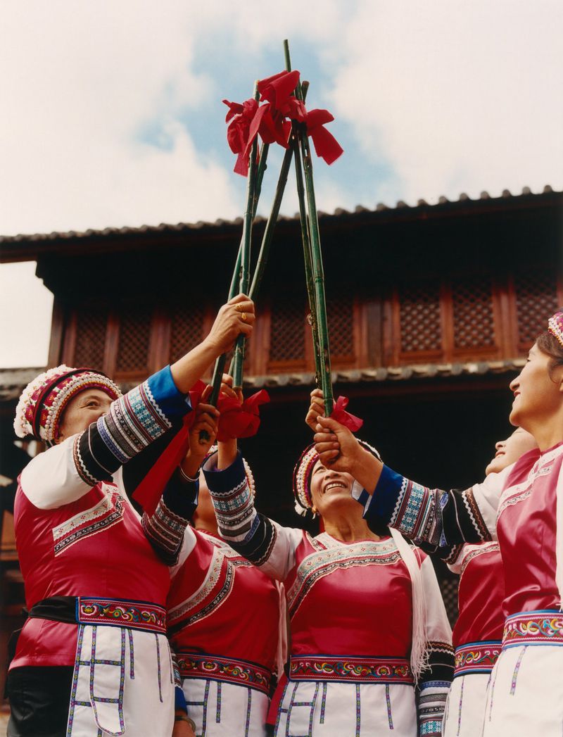 Group of women in traditional red clothing performing ceremony with bamboo sticks and red ribbons in outdoor setting with building.