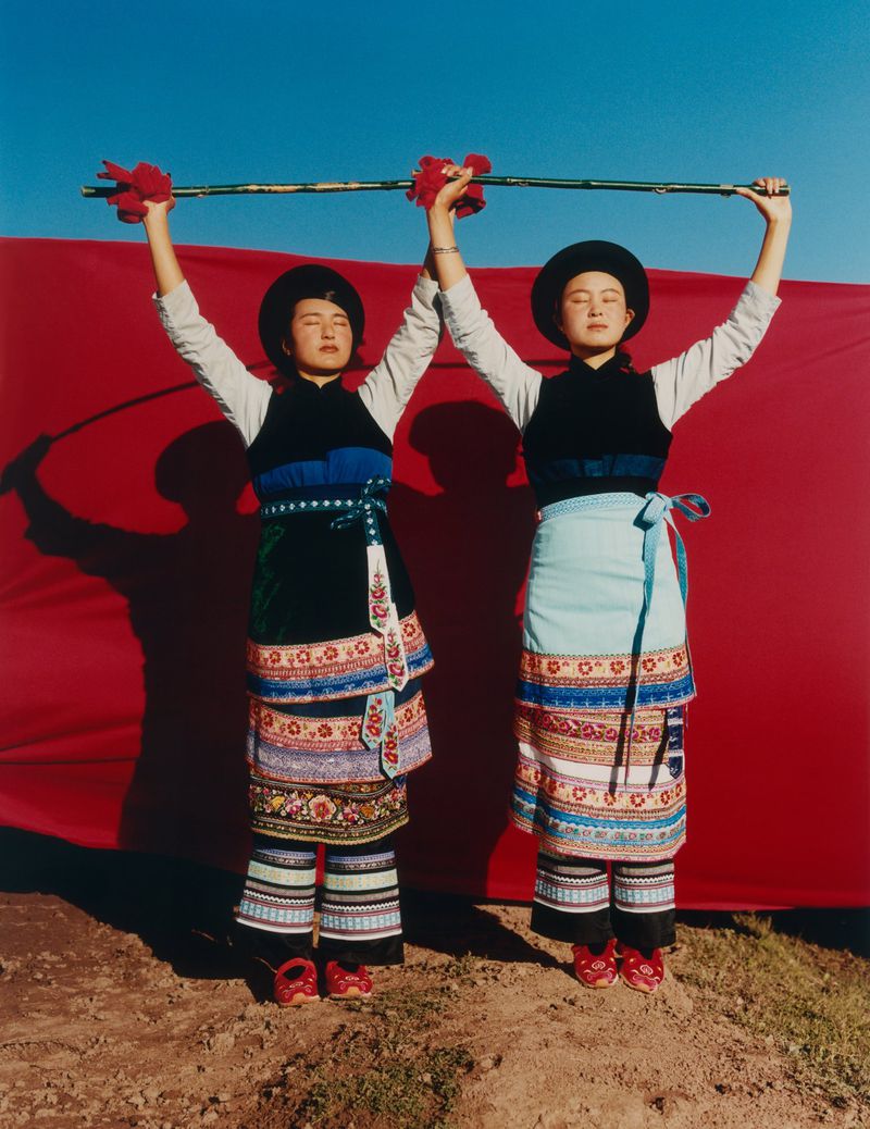 Two women in traditional clothing stand against a red backdrop under a blue sky, holding a bamboo pole decorated with red tassels.